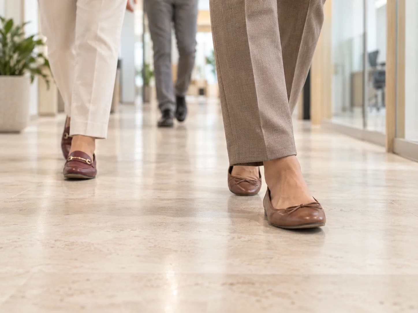 Close-up of anonymous workers' feet in motion across a bright office floor, with taupe trousers and burgundy shoes.